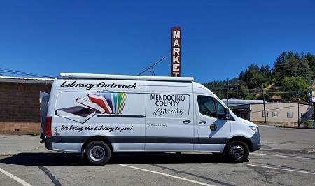 Outreach Van in a parking lot with a sign reading market in the background