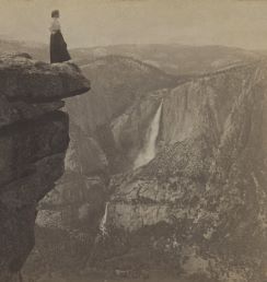 Black and white photo of a woman standing on a high cliff overlooking Yosemite, a waterfall can be seen in the distance. 