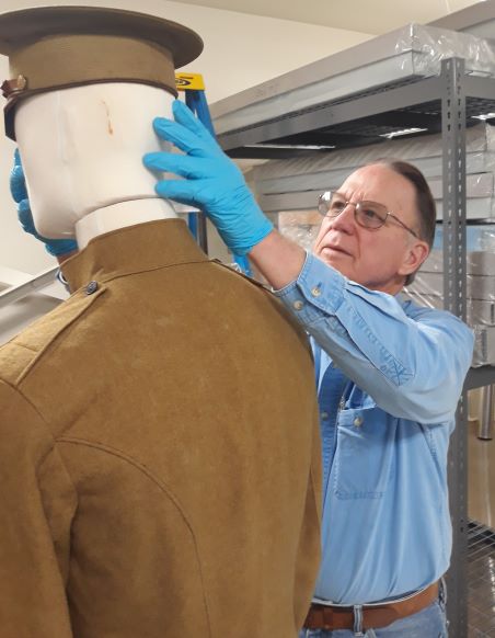 A man wearing a blue shirt adjusts the cap on a mannequin wearing a WWI uniform.  