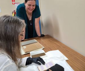 A woman sitting a table looks and documents, a second women looks towards the camera smiling.