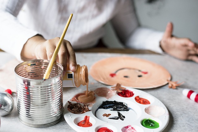 A child's hands are shown with paint on a palette, a painted smiley face, and a brush in a tin cup.