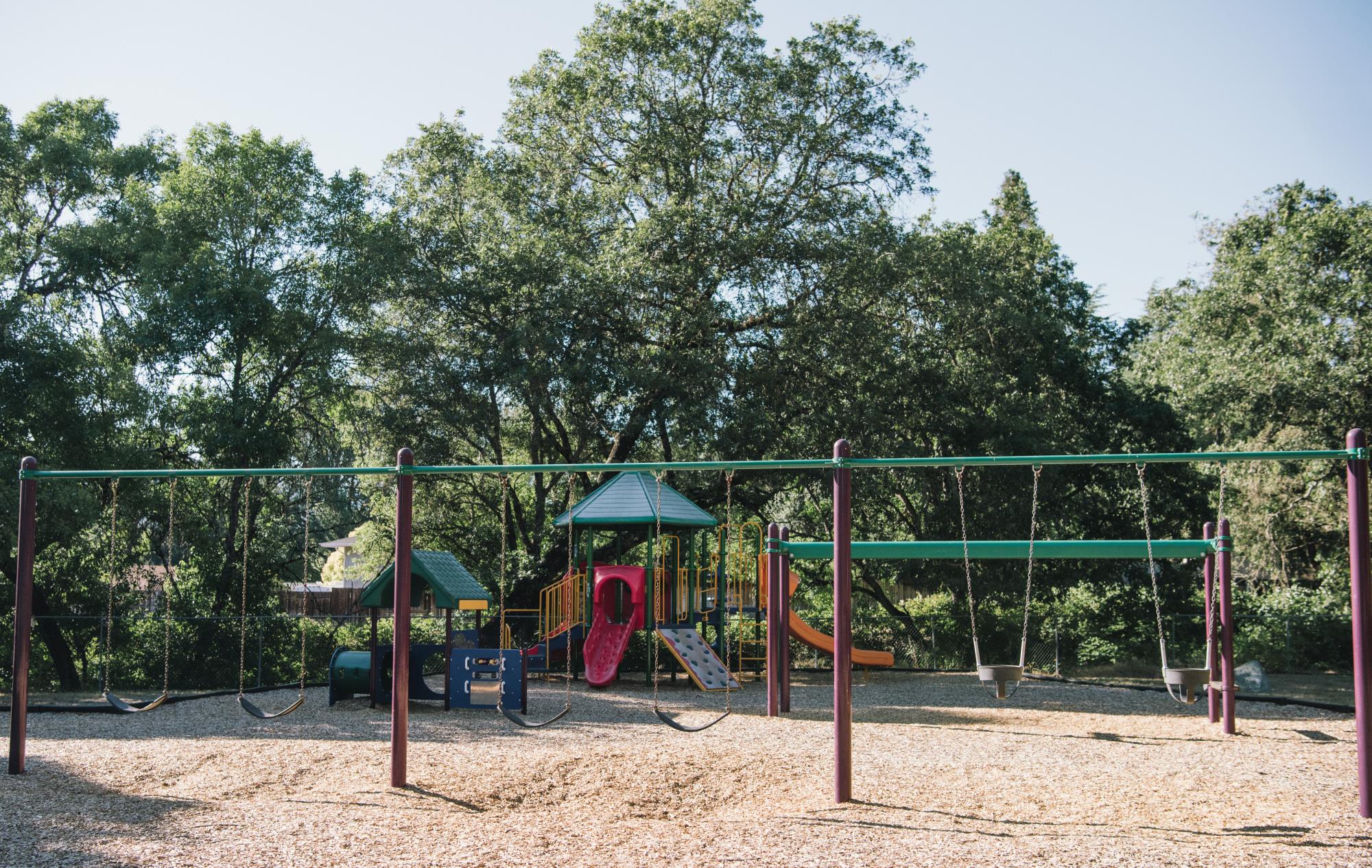 photo of the playground, including a swingset, a jungle gym, and slide set in woodchips